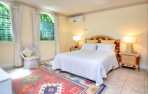 Un dormitorio ordenado con cama, dos mesitas de noche, una silla y un ventanal con cortinas.