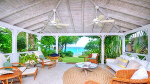 A covered outdoor seating area with wicker furniture, a central round table, and a view of the ocean. - Secret Cove 1 by Altman (Paynes Bay)