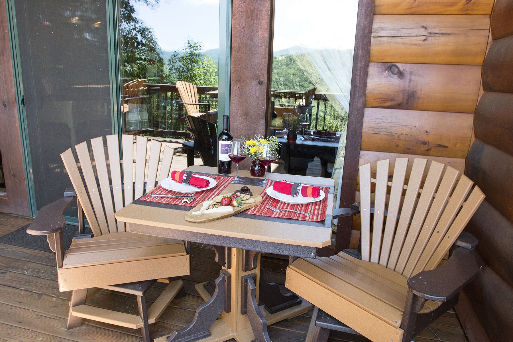 A wooden table with two chairs, a bottle of wine, and a glass of red wine on a deck with a view of mountains.