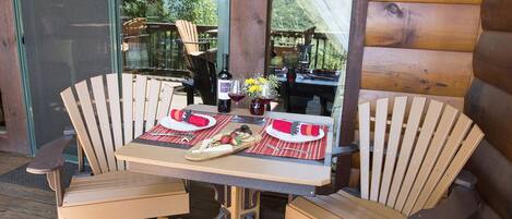 A wooden table with two chairs, a bottle of wine, and a glass of red wine on a deck with a view of mountains.