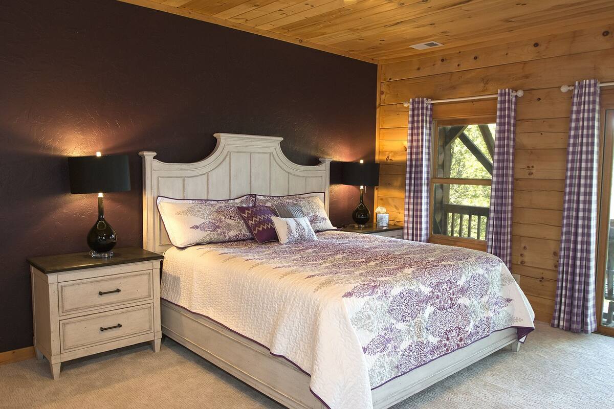 A bedroom with a wooden headboard bed, two bedside tables with lamps, a nightstand, and a window with curtains.
