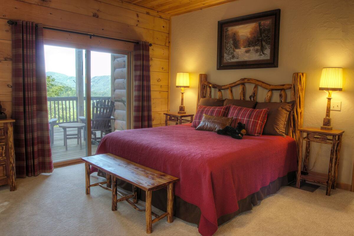 A cozy bedroom with a large bed, two bedside lamps, a wooden bench, and a view of the outdoors through a sliding door.