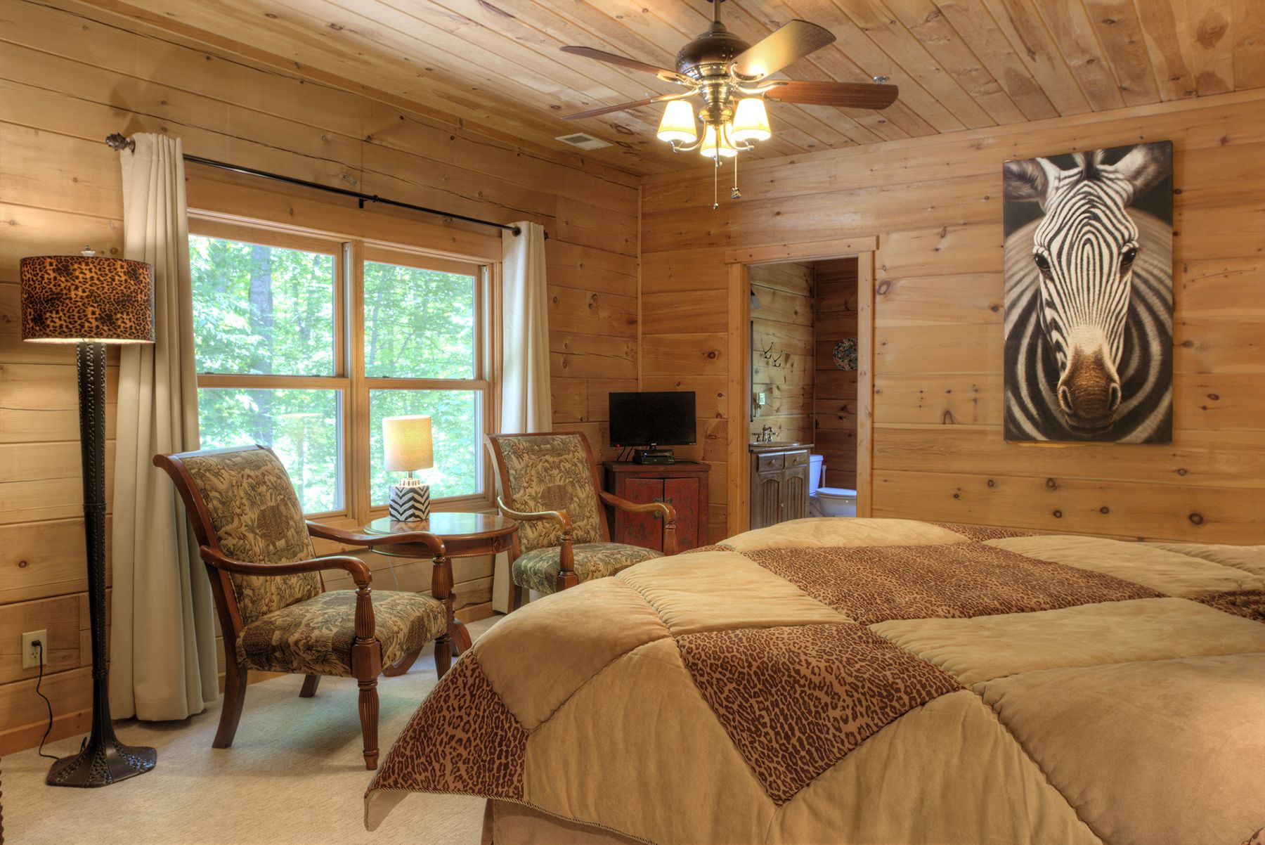 A wooden cabin room with a bed, two chairs, a table, a television, and a zebra painting on the wall.