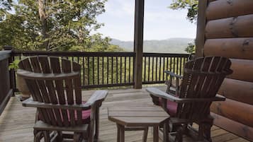 A wooden deck with two Adirondack chairs and a small table, overlooking a mountainous landscape.