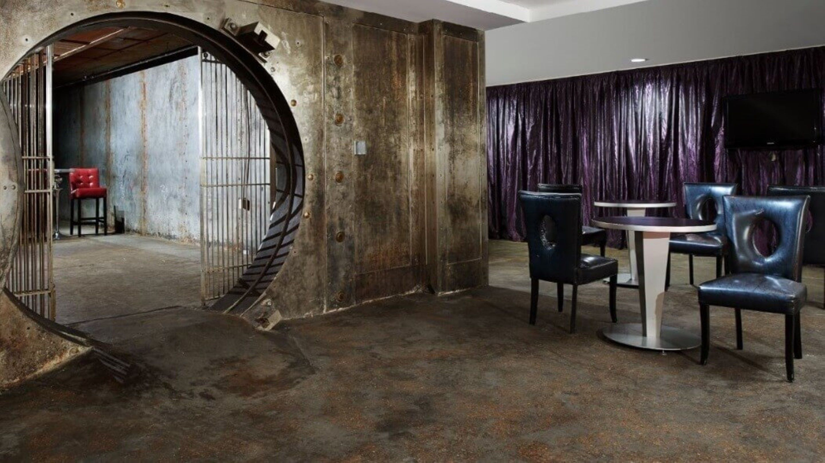 Una habitación con una puerta metálica circular, una mesa redonda con sillas y un telón de fondo morado.