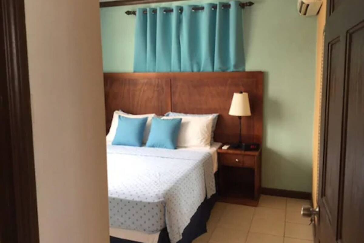 Una habitación de hotel con cama, mesita de noche, lámpara y ventana con cortinas.