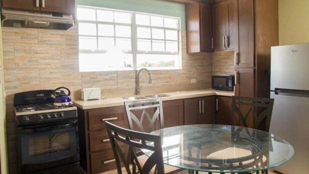 A kitchen with a gas stove, oven, microwave, and refrigerator. There is a window above the sink and a glass table with chairs. - Believe Caribbean Apartments (Bridgetown)