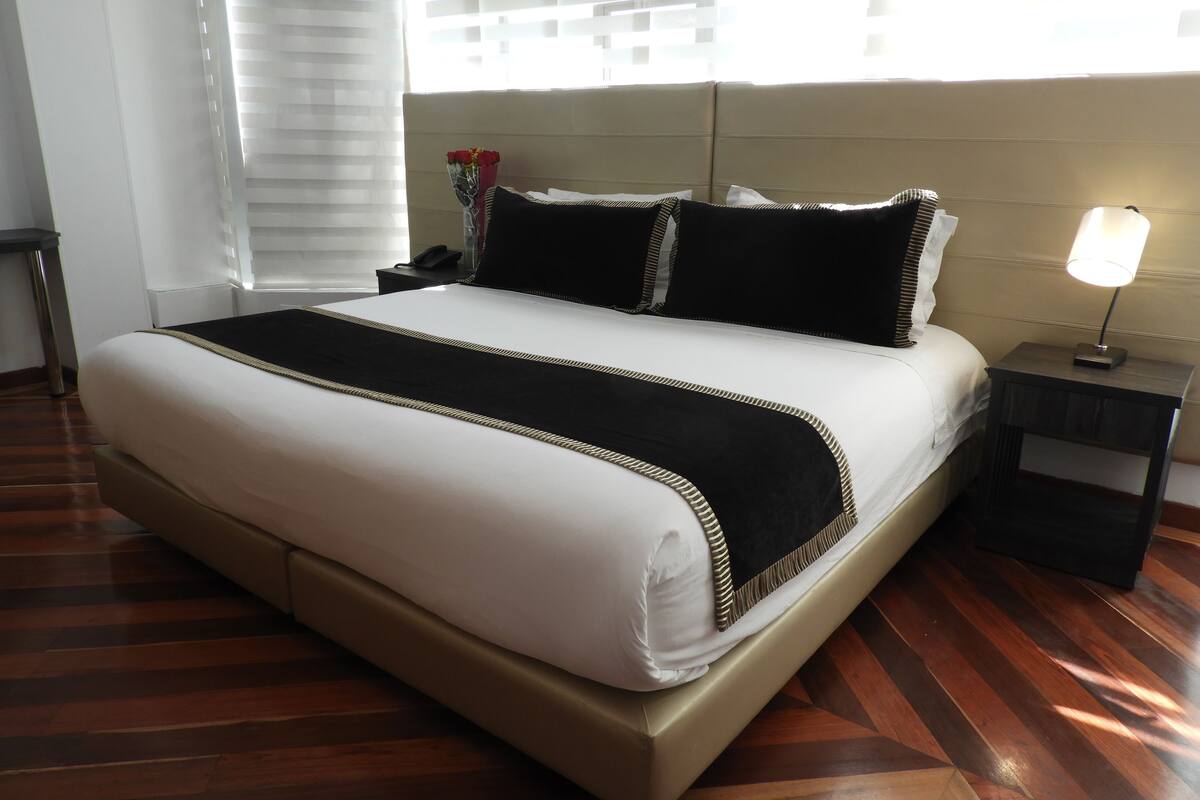 Una cama bien tendida con colcha blanca y negra, un detalle dorado y una almohada negra. Hay una lámpara de noche en una mesita de madera.
