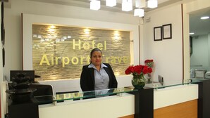 Una recepción con encimera de vidrio, un jarrón con flores rojas y una pared con el texto "Hotel Airport Travel".