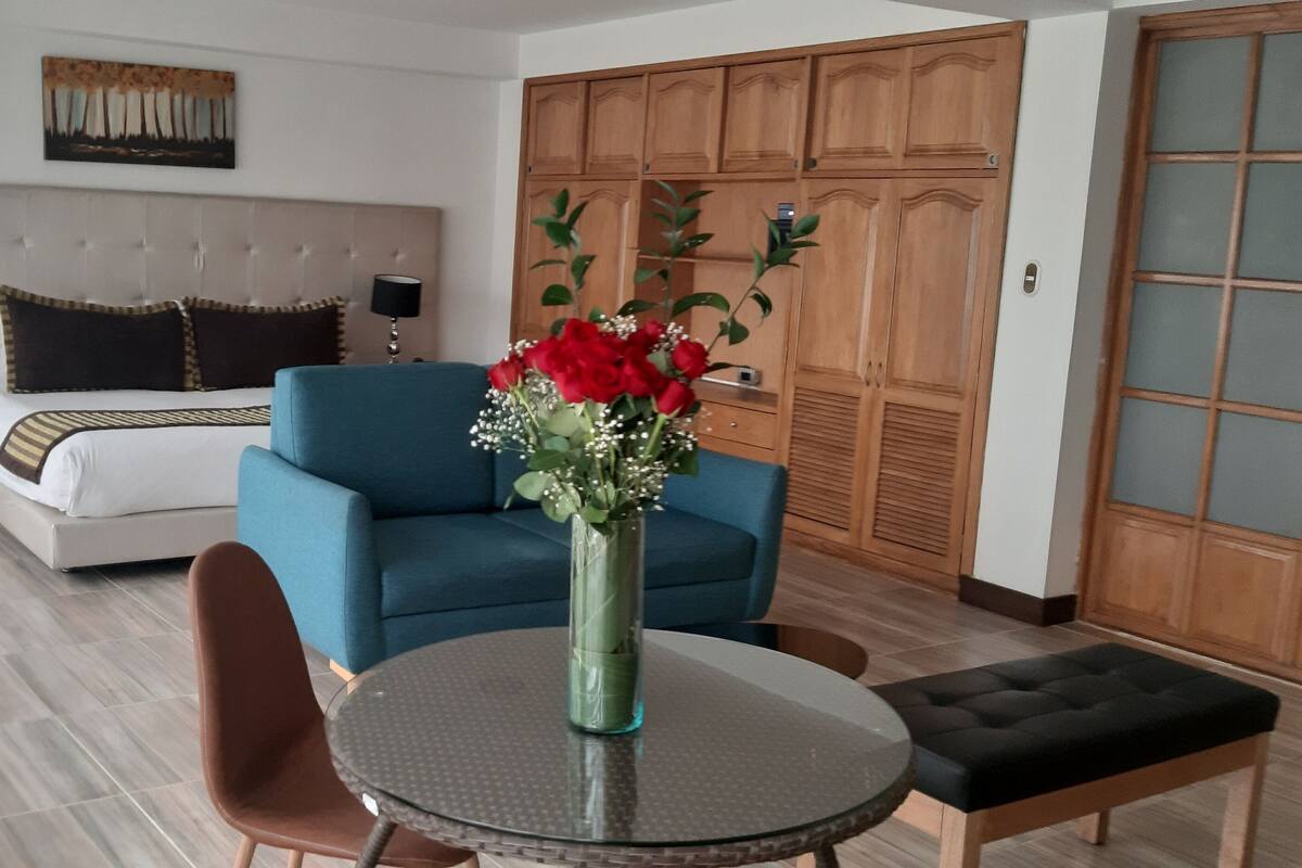 Habitación de hotel con una cama, un sofá azul, una mesa redonda con un florero y un armario de madera.