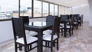 Un comedor moderno con mesas de vidrio y sillas oscuras, ventanas amplias con vista a la ciudad y suelo de baldosas.
