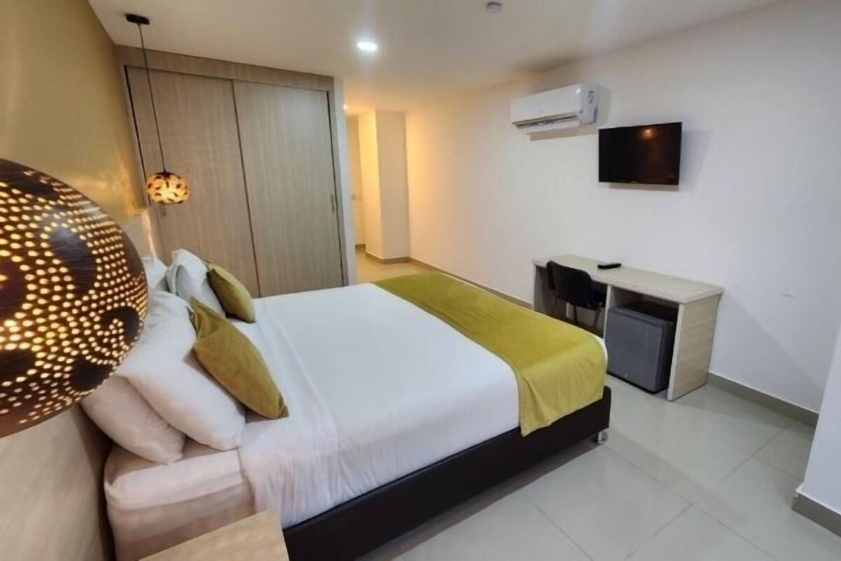 A hotel room with a bed, bedside table, desk, and an air conditioning unit.