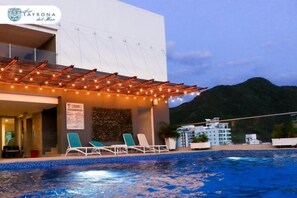 A hotel with a swimming pool, lounge chairs, and a mountain in the background. - Hotel Tayrona del Mar (Santa Marta)