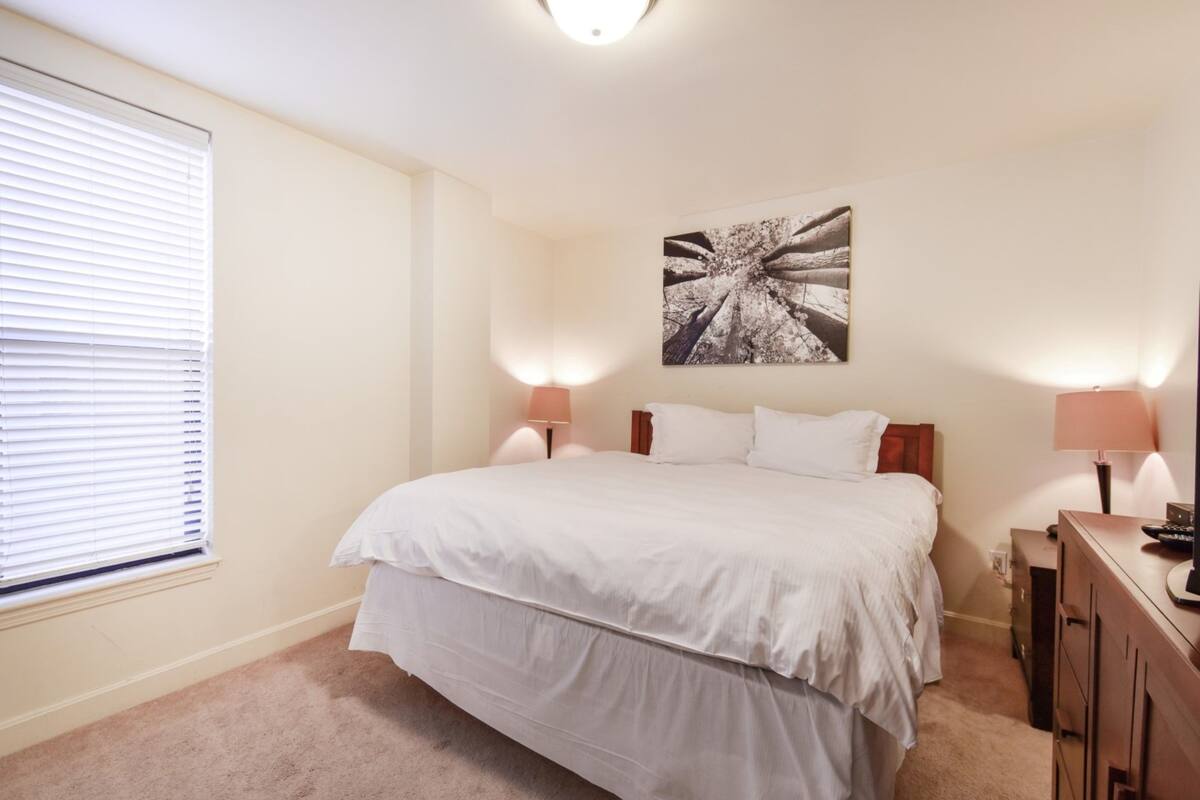 A bedroom with a large bed, two bedside lamps, a nightstand, and a wall-mounted artwork.