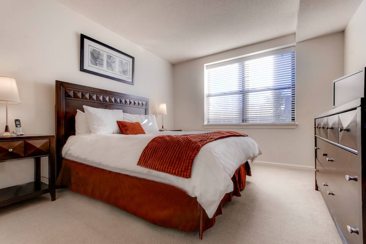 A hotel room with a large bed, a dresser, a nightstand, a window with blinds, and a framed picture on the wall.