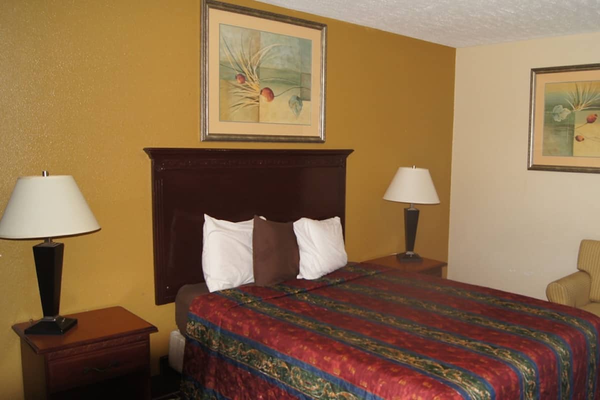 A hotel room with a large bed, two bedside lamps, a wooden headboard, a chair, and two framed artworks on the walls.