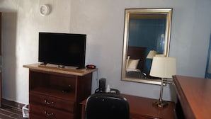 Habitación de hotel con una televisión sobre un tocador de madera, un espejo en la pared, una lámpara y una cama con cabecero.