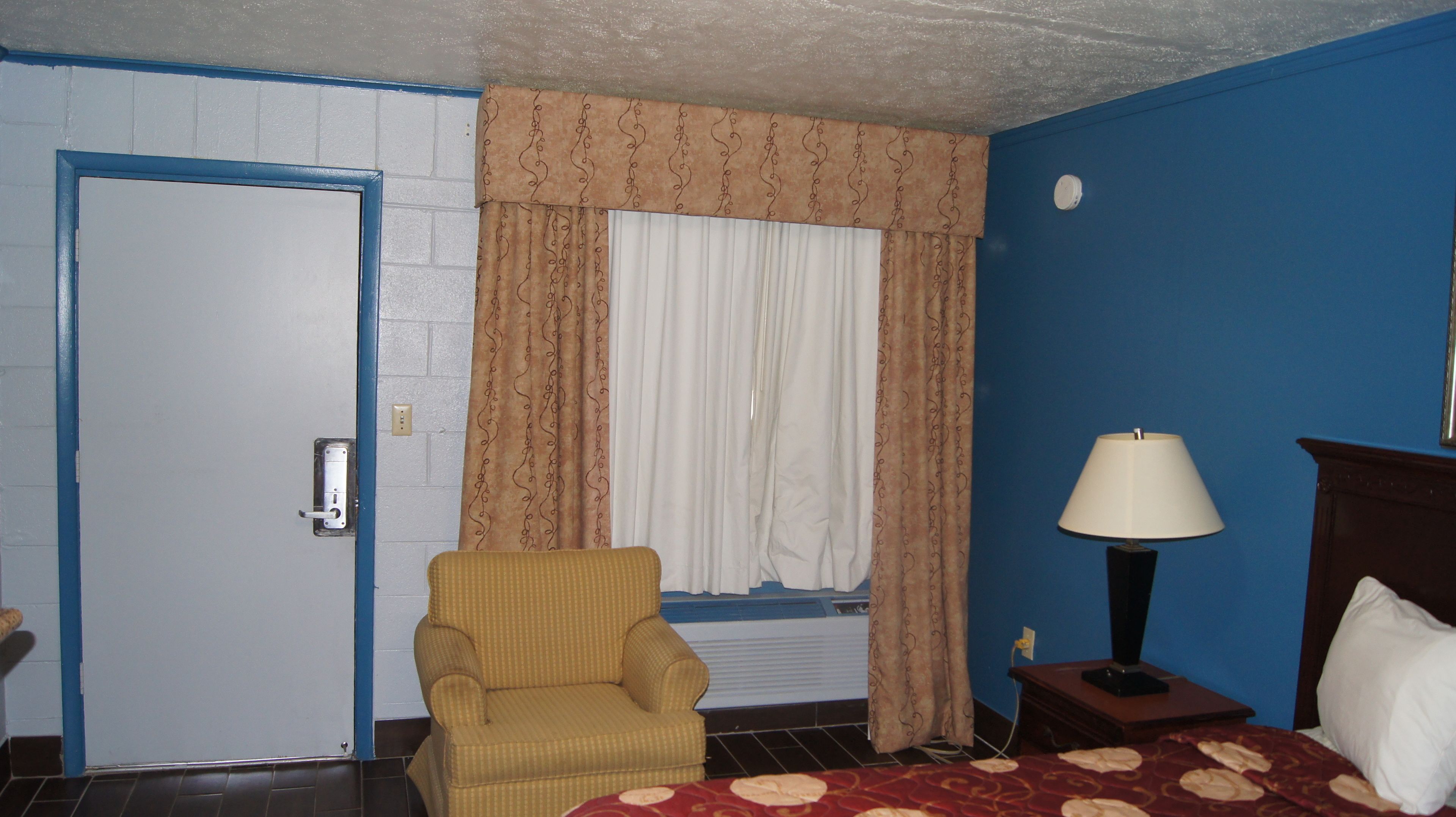 A room with a blue door, a yellow armchair, a bed with a red patterned bedspread, a bedside table with a lamp, and a window with curtains.