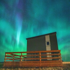 Una cabaña pequeña con terraza de madera, frente a un cielo nocturno estrellado y una aurora verde.