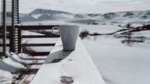 A white cup on a wooden bench with a snowy landscape and mountains in the background. - Afternoon Cottages (Rangárþing ytra)