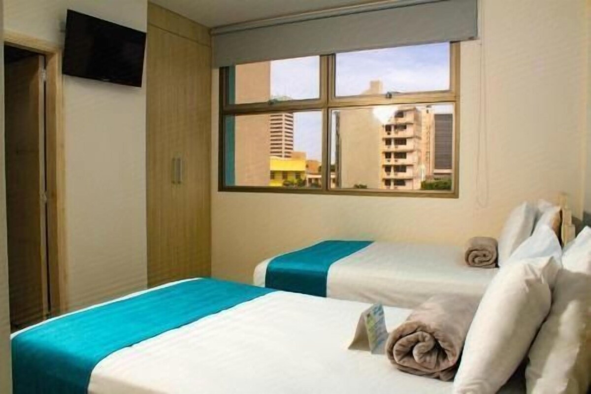 Habitación de hotel con dos camas, una ventana con vistas a edificios y un televisor en la pared.
