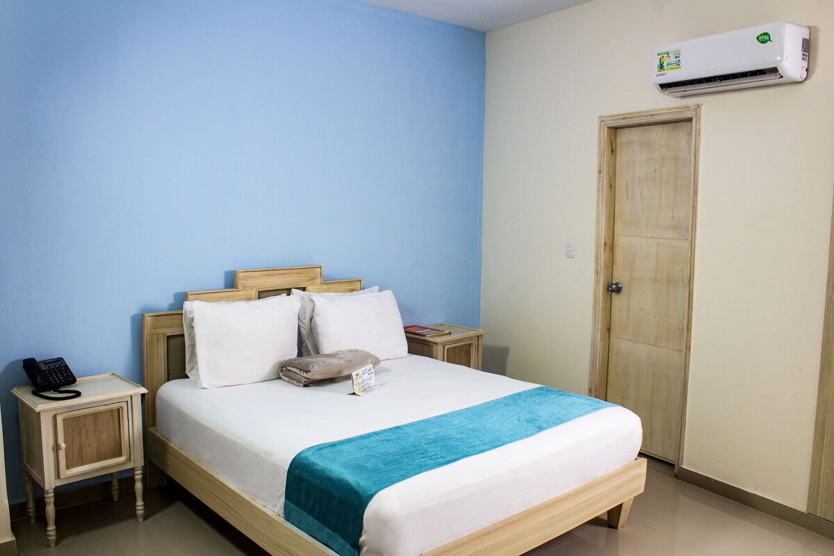 Habitación de hotel con cama, mesita de noche con teléfono, puerta de madera y unidad de aire acondicionado.