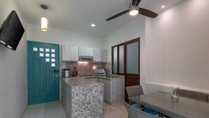 A compact kitchen with a teal door, white cabinets, a granite countertop, and a dining area with wicker chairs and a glass table. - Ka Suites Tulum by HA (Tulum)