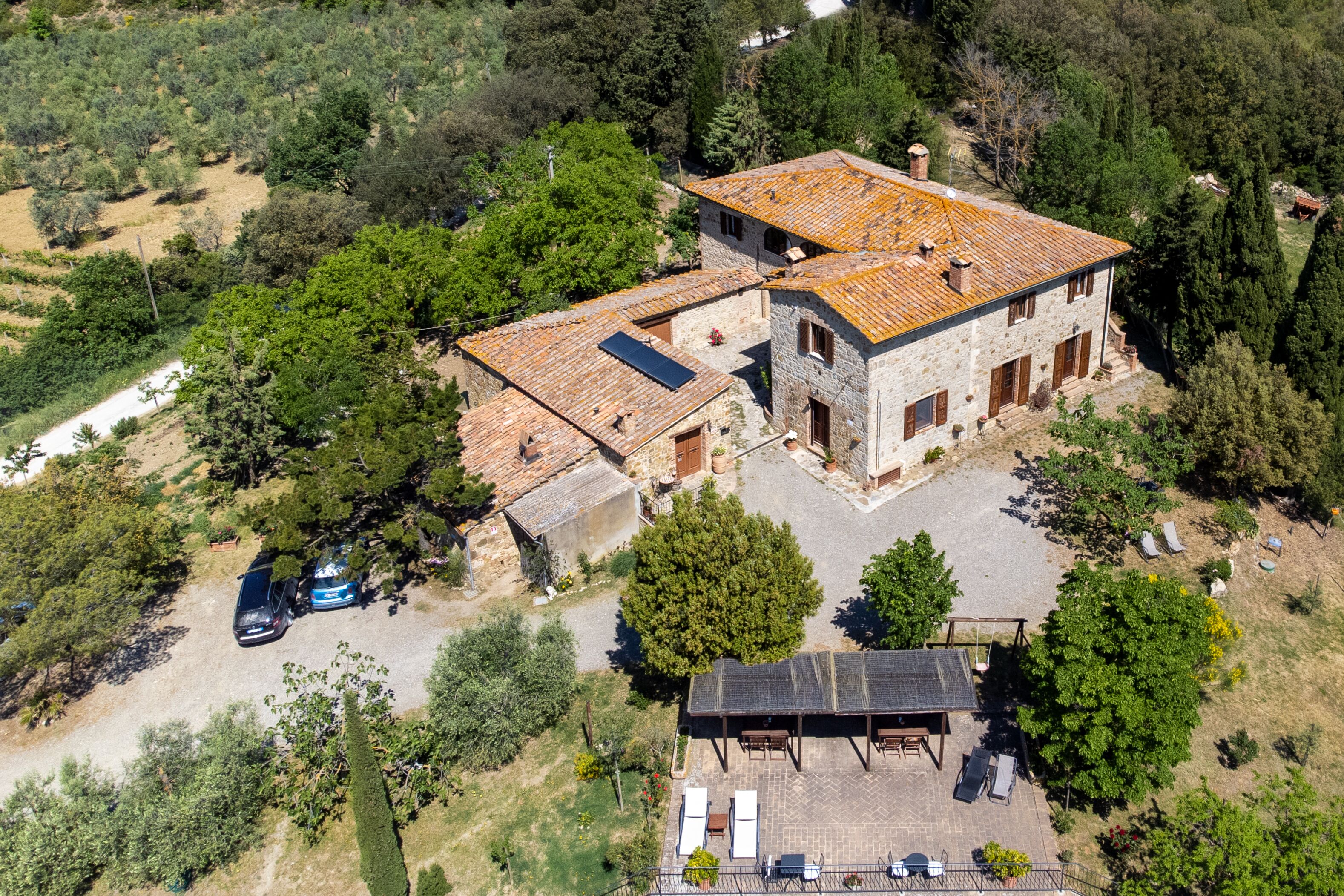 A two-story stone house with a tiled roof, surrounded by trees and a gravel driveway.