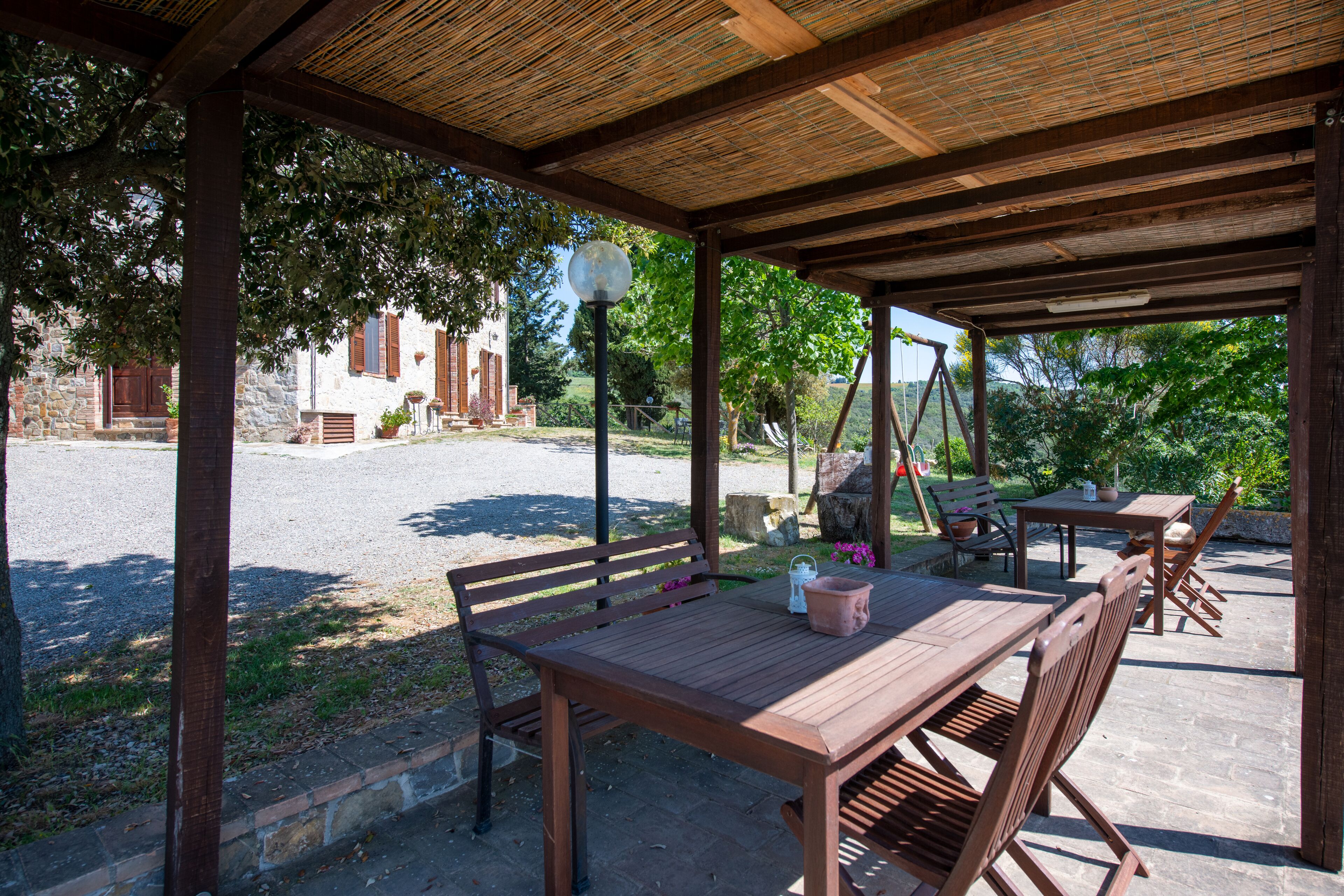 A covered outdoor seating area with wooden tables and chairs, a lamp post, and a view of a stone building in the background.