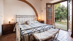 Un dormitorio con una cama, dos mesitas de noche, un banco, techo de madera, pared de ladrillo y una puerta que da a un jardín.