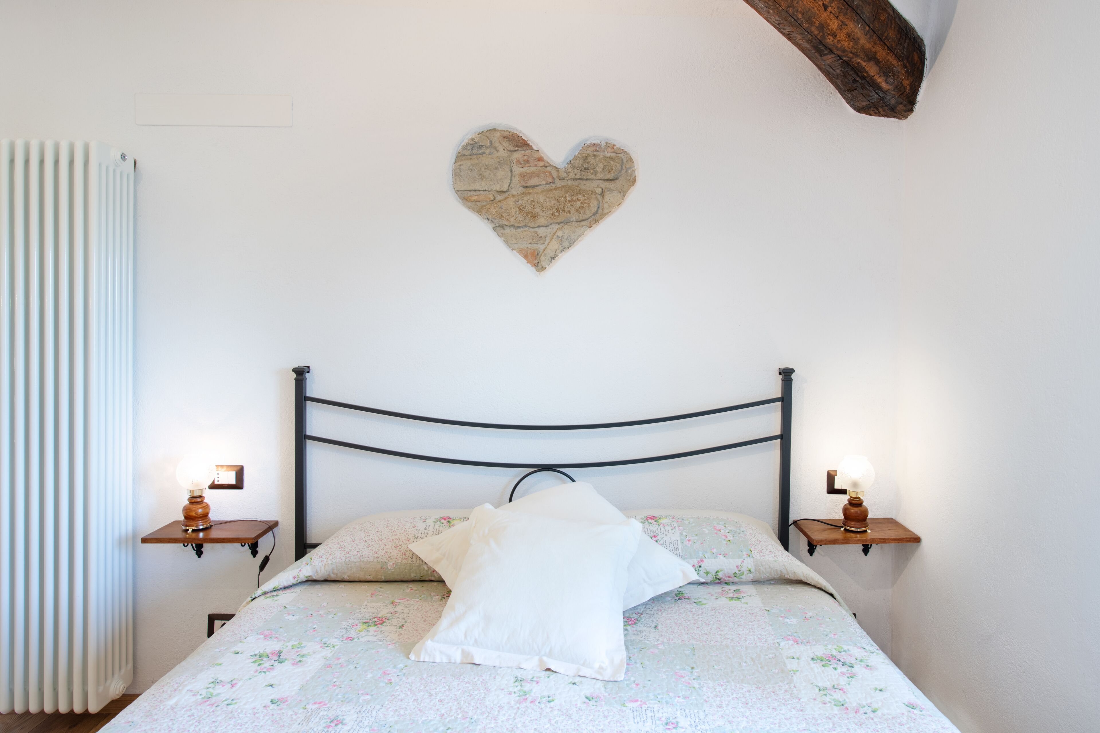 Un dormitorio con una cama, dos mesitas de noche, un adorno con forma de corazón en la pared y un radiador.