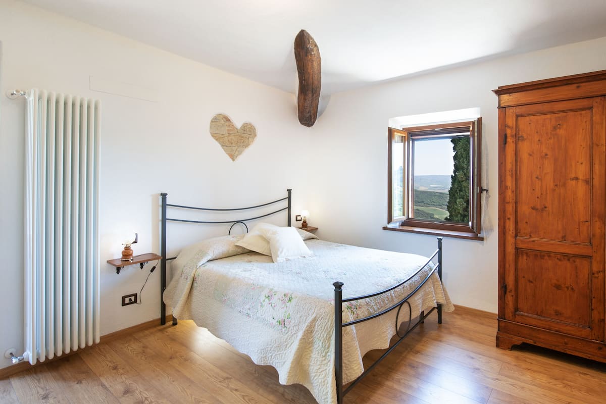 A bedroom with a bed, a wooden cabinet, a radiator, a window with a view, and a heart-shaped decoration on the wall.