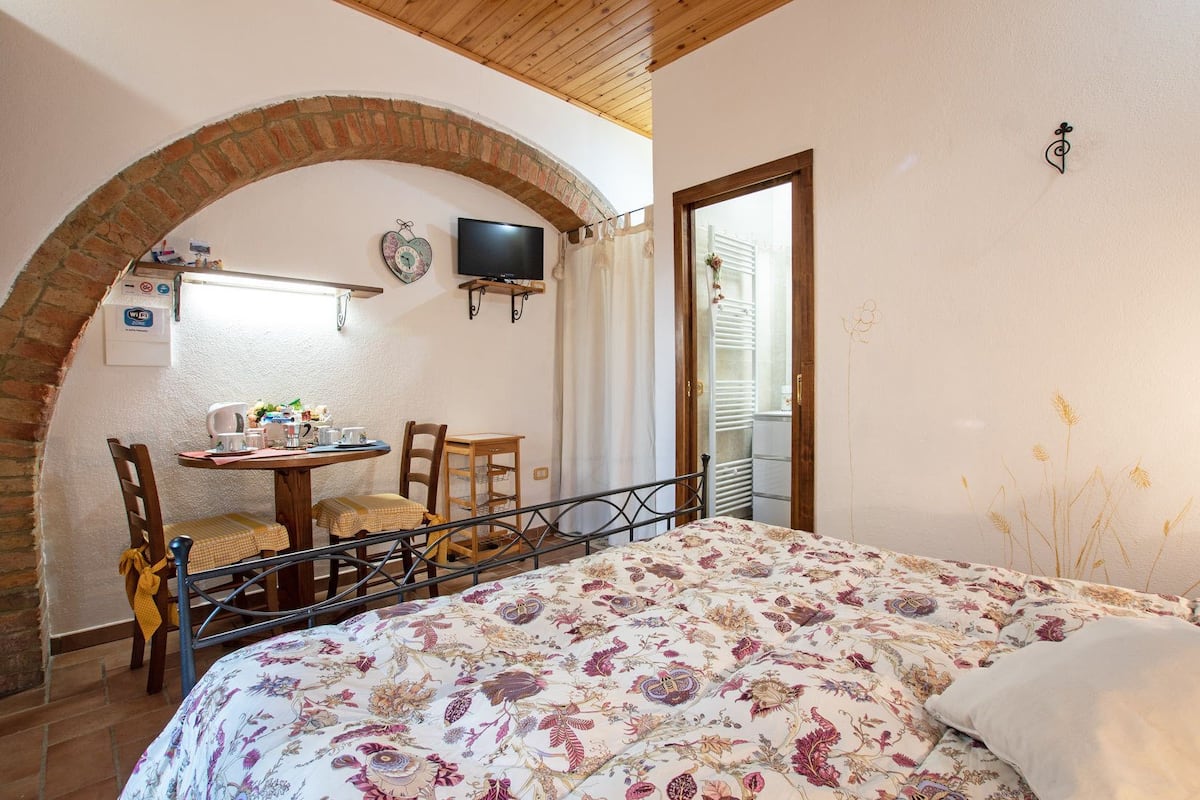 A cozy room with a bed, a table with chairs, a television, and a brick archway.