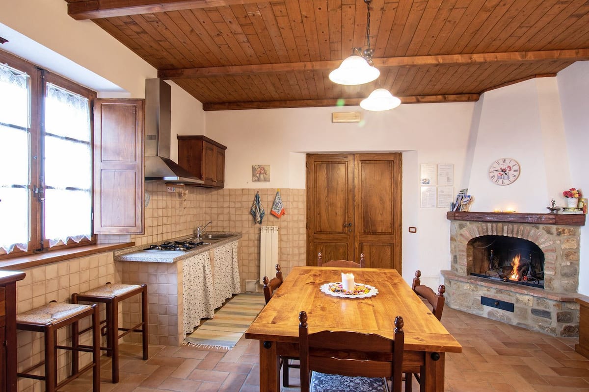 A cozy kitchen with wooden beams, a stone fireplace, and a dining table with benches.