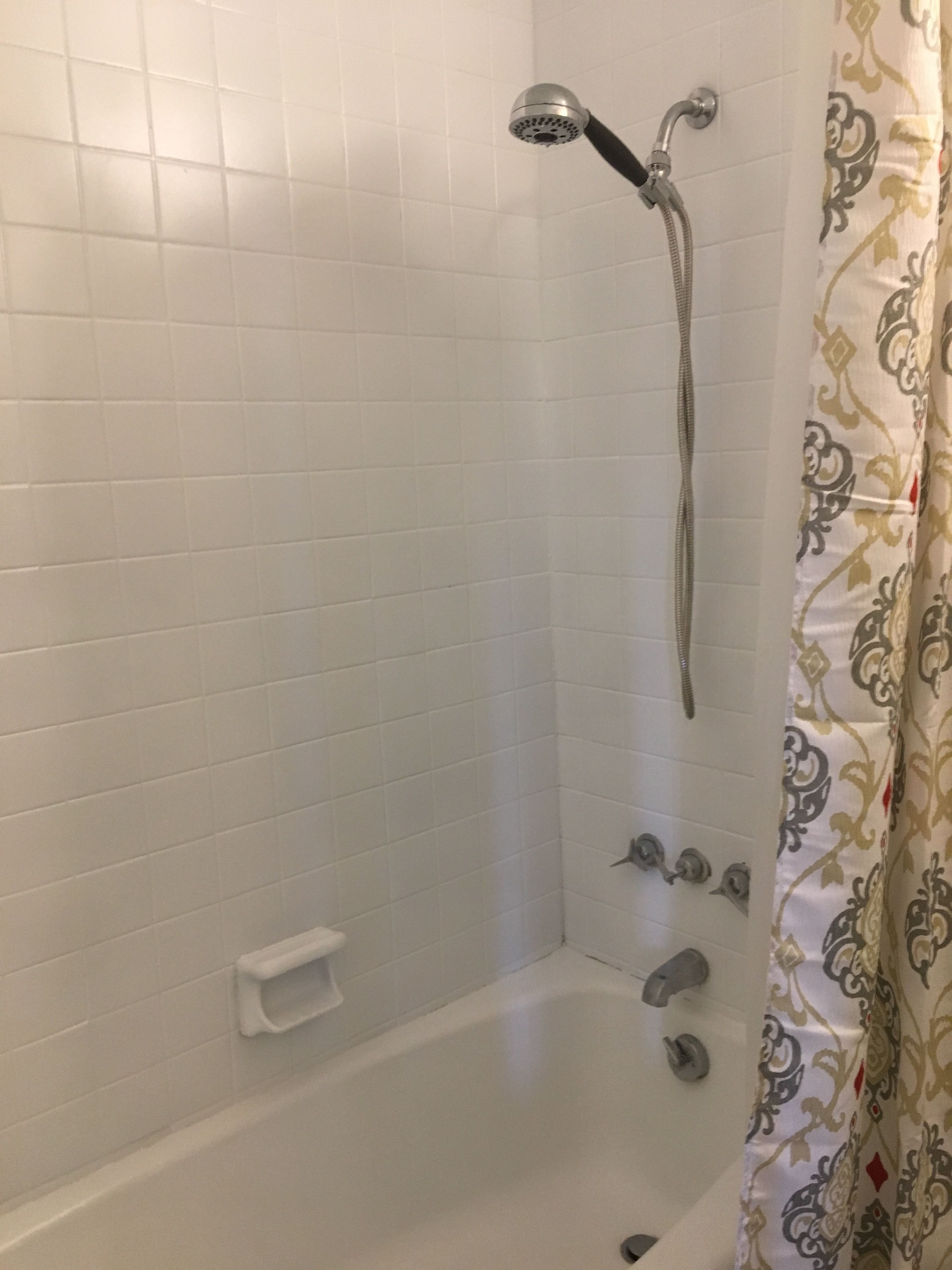 A bathroom with white tiled walls, a showerhead, a handheld shower, a small white shelf, and a patterned shower curtain.