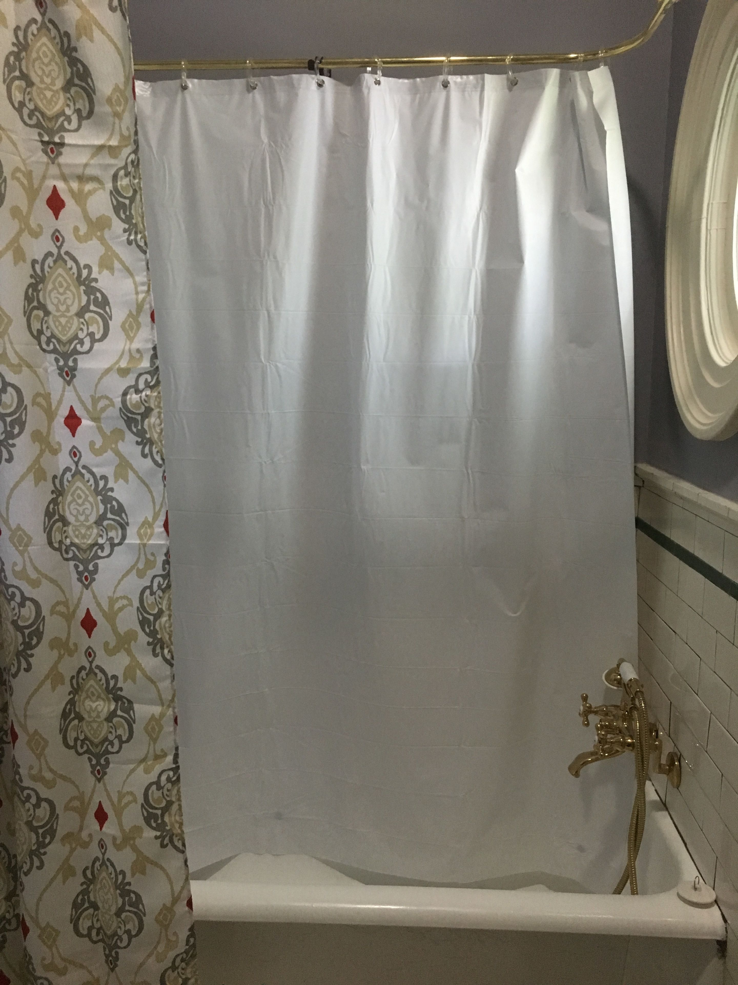 A bathroom with a white shower curtain, a gold showerhead, and a patterned curtain on the left.