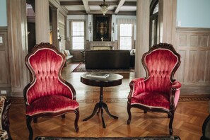 A room with two red upholstered chairs with wooden frames, a round wooden table, and a fireplace in the background. - 1912 Bed and Breakfast (Sumter)