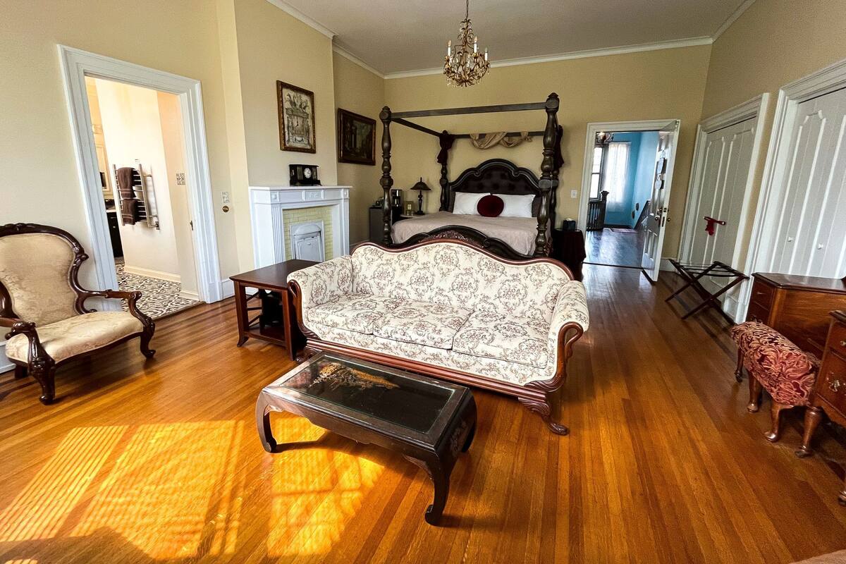 A spacious bedroom with a four-poster bed, a sitting area with a sofa and armchair, a fireplace, and a chandelier.