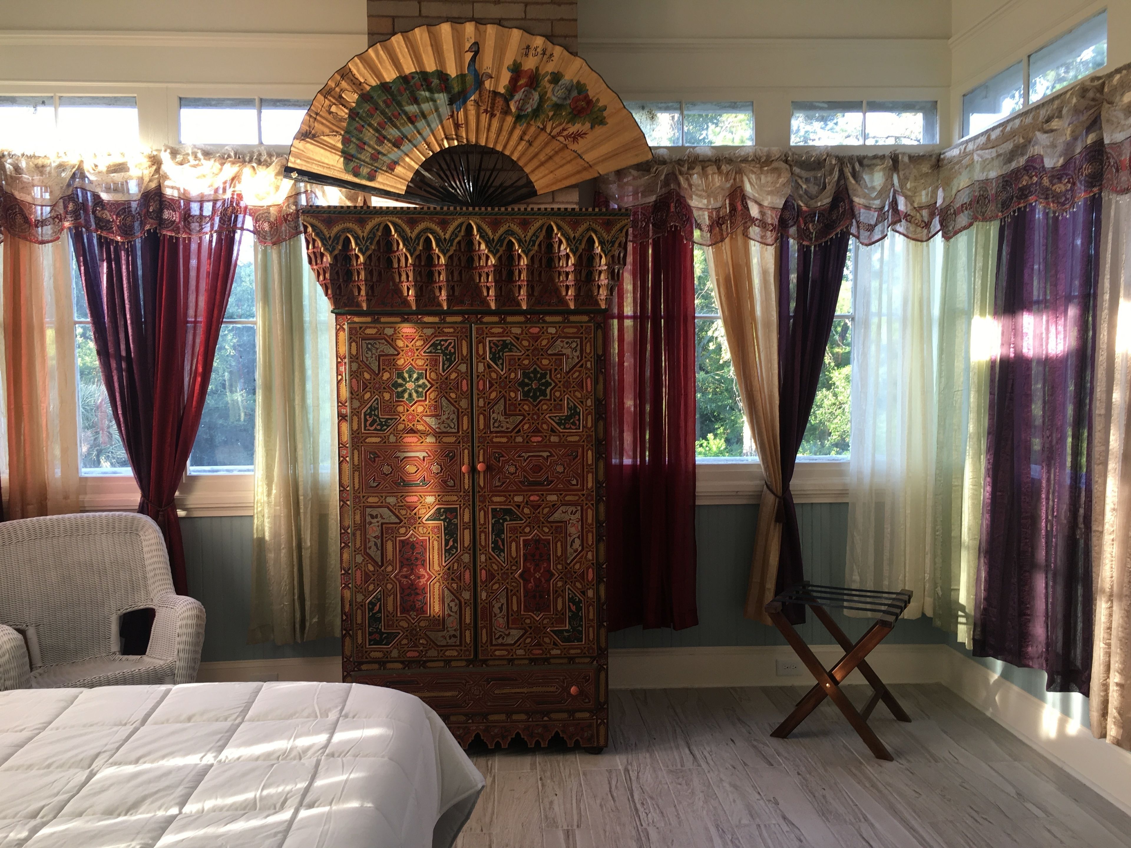 A room with a large bed, a wicker chair, a decorative cabinet, and a fan.