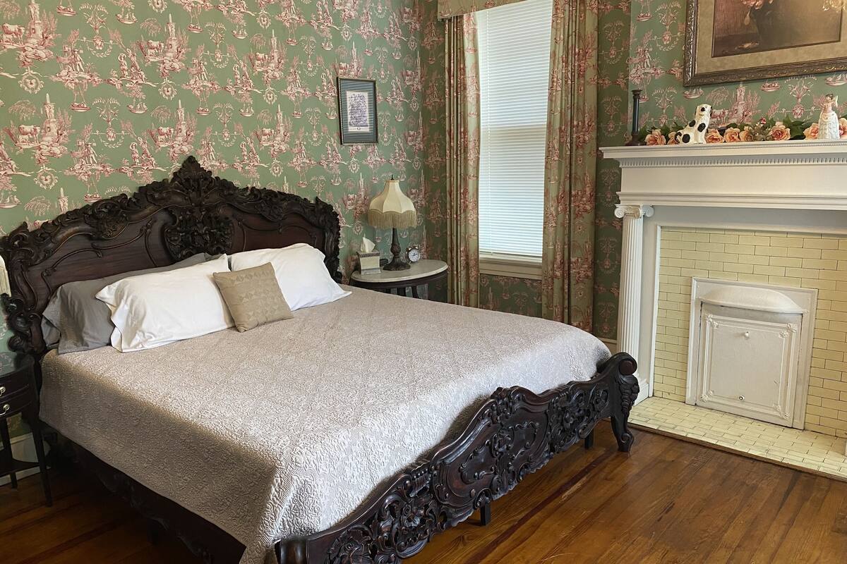 A bedroom with a large ornate bed, floral wallpaper, a fireplace, and a window with curtains.