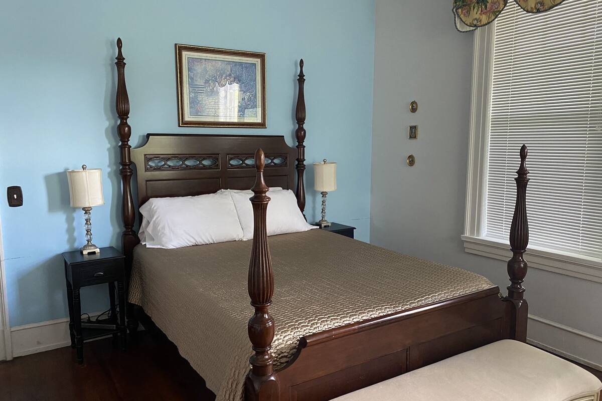 A bedroom with a wooden four-poster bed, two bedside tables with lamps, a framed picture on the wall, and a window with blinds.