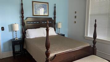 A bedroom with a wooden four-poster bed, two bedside tables with lamps, a framed picture on the wall, and a window with blinds.