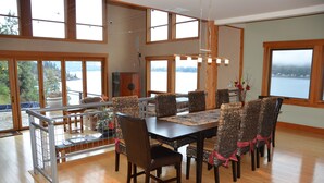 Un comedor con una mesa grande y sillas, ventanas amplias con vistas a un lago, y un balcón con barandilla.