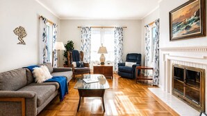 En stue med sofa, lænestole, et glasbord, pejs og mønstrede gardiner.