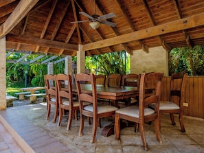 Un área de comedor al aire libre con techo, mesas y sillas de madera, un ventilador de techo y vistas a vegetación exuberante.