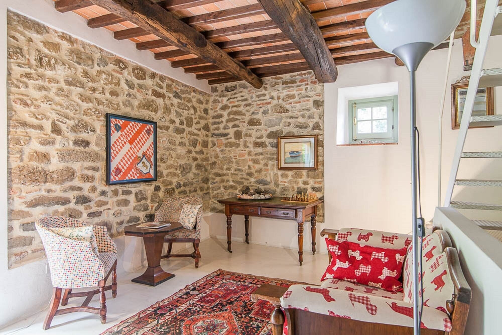 A cozy room with a stone wall, wooden beams, a patterned rug, a rocking chair, a small table with a chessboard, and a sofa with patterned cushions.
