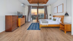 A bedroom with a wooden ceiling, a bed with pillows, a nightstand, a dresser, a TV, and a view of the sunset through the window.