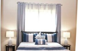 A neatly made bed with a striped bedspread, two bedside tables with lamps, and a window with curtains. - "The Indigo" A little bit country, close to town.  New, clean and fresh looking (Kyle)