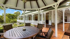 Un espace extérieur aménagé avec une zone salon couverte, comprenant une table ronde en bois et des chaises, le tout entouré de rampes blanches et offrant une vue sur une végétation luxuriante.