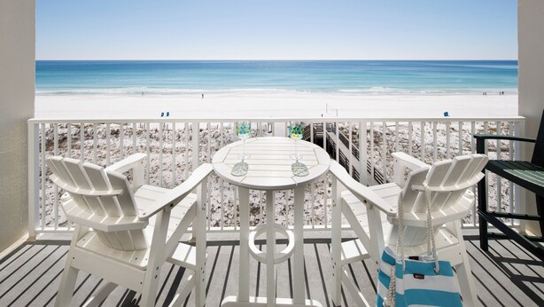 A balcony with a view of a beach, white chairs, and a table.
